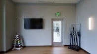 This single-floor space appears to be a fitness or activity room designed with functionality and simplicity in mind. The room features neutral gray walls and light wood-tone flooring, creating a clean and modern aesthetic. A large flat-screen television is wall-mounted on one side, serving as a focal point for entertainment or guided workout sessions. To the right, a vertical equipment rack holds barbells or similar exercise tools, indicating the room's intended use as a workout or training area. A glass-paneled door with an illuminated exit sign above it provides both a safety egress and a view into an adjacent space, which appears to be a brighter, open area — possibly a lounge or common zone. The door's glass panel allows natural light to filter into the room, adding a sense of openness. Ventilation is provided through a ceiling vent, contributing to a comfortable environment during physical activity. The overall design is minimal and utilitarian, prioritizing practical use while maintaining a tidy, well-organized appearance suitable for group or individual fitness activities.