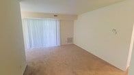 This single-floor space presents a straightforward, functional layout centered around a generously sized living area finished with wall-to-wall beige carpeting that adds warmth underfoot. The room is characterized by clean, cream-colored walls that provide a neutral canvas adaptable to a variety of interior styles. A prominent sliding glass door dominates one wall, dressed with vertical blinds that allow for adjustable light control while offering access to the outdoors. Natural light filters in through the covered blinds, contributing to a bright and airy atmosphere within the room. A wall-mounted HVAC vent provides climate control, and a ceiling-mounted ventilation register further supports air circulation throughout the space. The overall design is minimal and unfussy, with smooth wall surfaces and standard ceiling height contributing to a tidy, open feel. The absence of furnishings highlights the versatility of the floor plan, allowing prospective occupants to envision a variety of configurations. This space offers a clean, move-in-ready foundation suited for comfortable everyday living.