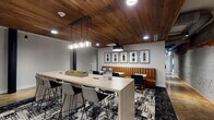 Located at 701 Easley Bridge Rd in Greenville, SC, this single-floor space masterfully combines industrial heritage with contemporary design. The layout flows through several distinct zones, each with its own character while maintaining a cohesive aesthetic throughout.

The main gathering area features rich wood-planked ceilings that contrast beautifully with polished concrete floors in warm amber tones. A central kitchen bar with dark textured paneling and bold blue bar seating anchors one section, equipped with stainless steel appliances and dark cabinetry beneath pendant lighting. Exposed ductwork and high clerestory windows allow natural light to filter in along the original brick walls, preserving the building's industrial roots.

A sophisticated lounge area showcases a wall-mounted electric fireplace framed by a textured dark surround and wood shelf, complemented by plush green velvet seating, an ornate chandelier, and a patterned area rug. The dining zones offer a mix of butcher-block communal tables with black chairs and upholstered leather banquette seating paired with navy accent chairs. Curated artwork and sculptural pendant lighting add refined detail throughout, creating a versatile, welcoming environment suitable for collaboration or relaxation.