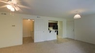 This single-floor space presents a straightforward, functional layout centered around an open living area with neutral white walls and wall-to-wall carpeting. The living space connects visually to the kitchen through a built-in pass-through counter with a breakfast bar overhang, creating a sense of openness while maintaining a defined separation between areas. The kitchen, visible through the pass-through, features dark cabinetry, a black refrigerator, and light countertops, with tile or hard flooring distinguishing it from the carpeted living area. A ceiling-mounted HVAC vent is visible, indicating a central air system. The space includes multiple doorways — one leading to what appears to be a hallway or additional rooms with warm lighting visible beyond, and another serving as the main entry door. The white six-panel interior doors contribute to the clean, uniform aesthetic throughout. Overall, the space is practical and uncluttered, with a neutral color palette that offers flexibility for various furnishing styles. The layout makes efficient use of the available footprint, with the pass-through kitchen serving as a functional design highlight.