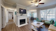 This thoughtfully designed single-floor residence offers a functional and comfortable layout across one bedroom and two full bathrooms. The open-concept living, dining, and kitchen area forms the heart of the home, featuring durable hardwood-style flooring, crown molding, and ceiling fans throughout. The living room is anchored by a classic white fireplace surround with a mounted television above, while large windows invite an abundance of natural light. A dedicated dining area adjacent to the kitchen benefits from oversized windows and a glass-panel door that opens to the outside, enhancing the bright and airy atmosphere. The galley-style kitchen is equipped with light wood cabinetry, granite-style countertops, a double sink, a black appliance suite including a range and dishwasher, and tile flooring. The spacious primary bedroom features plush carpeting, a ceiling fan, and generous natural light from a large window. The two bathrooms include tile flooring, wood-tone vanity cabinetry, and a soaking tub with decorative tile accents. A dedicated laundry room with full-size washer and dryer hookups and built-in shelving adds everyday convenience. A hallway and closet complete the practical and well-organized floor plan.