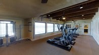 This fitness center offers a dedicated and thoughtfully designed workout environment ideal for cardiovascular training. The space is defined by its distinctive exposed wooden beam ceiling, which adds a warm, rustic character to the room while ceiling fans ensure comfortable air circulation throughout exercise sessions. A row of elliptical cross-trainers lines the space, all positioned to face a series of large windows that span the exterior wall. These windows allow abundant natural light to pour in, creating an open and energizing atmosphere. The walls feature a subtle geometric blue-framed decorative pattern, adding a modern design touch to the room. The flooring consists of protective rubber mats beneath the equipment, transitioning to a durable, easy-to-maintain floor surface throughout the rest of the space. Additional cardio equipment, including treadmills, can be seen extending toward the back of the room. The overall ambiance is functional yet inviting, balancing practical fitness amenities with architectural details that make the experience of working out more engaging and visually appealing.