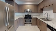 This single-floor residence is designed with a clean, contemporary aesthetic that carries consistently through each space. The kitchen is a standout feature, outfitted with flat-front cabinetry in a warm taupe wood-grain finish, light speckled granite countertops, and a full suite of stainless steel appliances, including a gas range with overhead ventilation, a French-door refrigerator, and a built-in microwave. Large-format tile flooring and a built-in counter nook add both practicality and style to the kitchen area.

The open layout connects the kitchen to a living area finished with warm wood-tone flooring, providing a cohesive and inviting atmosphere. Multiple doors lead to the bedrooms, one of which features soft carpeting and a window with an exterior view, allowing natural light to fill the room.

The unit includes two bathrooms, both finished in a bright, minimalist style with white walls, light tile flooring, and light wood vanity cabinetry paired with white countertops and frameless mirrors. One bathroom offers an extended vanity with additional storage, while the other features a compact single-sink vanity with clean chrome fixtures. Both spaces convey a sense of calm functionality consistent with the overall design of the residence.