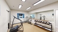 Located at 1201 Edenham Ct in Virginia Beach, VA, this fitness room is a well-equipped exercise facility designed to support a range of workout routines. The single-room space is finished with neutral-toned carpet flooring and crisp white walls, creating a clean and functional environment. Overhead, rectangular flush-mount fluorescent light panels provide consistent, bright illumination throughout the room.

A defining architectural feature is the large wall-mounted mirror spanning one side of the room, paired with a wood-tone ballet barre — enhancing both the sense of space and the functionality of the room for exercise and stretching. A pass-through interior window on the adjacent wall offers a view into a neighboring common area, adding visual interest and openness.

The room is equipped with a variety of cardio and strength training equipment, including a treadmill, stair climber, stationary cycle, air resistance bike, and a multi-function cable machine. A dumbbell rack stocked with free weights provides additional strength training options. A dedicated exit door with full-length glass panel allows natural light to enter and provides convenient outdoor access. Posted exercise room rules and a hand sanitizer dispenser reflect a commitment to cleanliness and safety. Overall, the space offers a practical and accessible fitness environment.