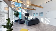 This single-floor space showcases a thoughtfully designed open lounge area that combines modern aesthetics with warm architectural details. The most striking feature is the soaring ceiling height, accented by exposed natural wood beams that add warmth and visual structure to the otherwise crisp, white interior. Large floor-to-ceiling windows along the exterior wall allow abundant natural light to pour in throughout the day, creating an airy and welcoming atmosphere.

The flooring throughout is light wood-tone laminate or hardwood, contributing to the space's clean, contemporary feel. A central seating arrangement features charcoal-gray sectional and lounge sofas oriented around a wall-mounted flat-screen television and a tiled fireplace, creating a comfortable focal point for the room. Decorative wall art, including colorful circular accents, adds a playful touch to the neutral palette.

Overhead, distinctive spherical pendant lights with slatted white shades serve as both functional lighting and sculptural design elements. Lush indoor plants, including a prominent fiddle-leaf fig, introduce organic texture and life to the space. Additional seating areas with teal accent chairs and a yellow ottoman provide flexible, vibrant touches throughout.