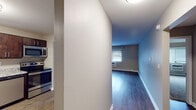This single-floor residence offers a clean, functional layout designed for comfortable everyday living. The open-concept kitchen is a standout feature, showcasing rich dark wood cabinetry with sleek hardware, granite-look countertops, a stainless steel sink, and a full suite of stainless steel appliances including a range, over-the-range microwave, and dishwasher. A breakfast bar with overhang connects the kitchen to the main living area, making the space ideal for both cooking and entertaining. The living area is generously proportioned, featuring dark hardwood-style flooring, neutral warm-toned walls, white baseboard trim, and a window with blinds that allows natural light to fill the room. A wall-unit air conditioner and baseboard heating provide year-round climate control. The bedrooms are carpeted for comfort and include windows with blinds for natural light. The primary bedroom features a double-door closet offering ample storage. Additional rooms include a smaller bedroom or flex space, also carpeted with a window. The bathroom is finished with wood-look flooring, a dark wood vanity, white countertop, and a tiled shower surround. Throughout the space, consistent neutral tones and wood accents create a cohesive, understated aesthetic.
