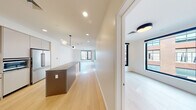 This thoughtfully designed single-floor residence combines clean, contemporary architecture with a bright, airy aesthetic throughout. Light hardwood flooring extends seamlessly from the entry hallway through the living areas and bedrooms, creating a cohesive sense of flow and warmth.

The long, open hallway leads into the heart of the home, where a well-appointed kitchen takes center stage. The kitchen features a mix of warm wood and white cabinetry, a white tile backsplash, stainless steel appliances including a large French door refrigerator, a built-in oven, and a spacious dark-topped island that provides ample prep and dining space. Globe pendant lighting adds a modern, sculptural touch overhead.

The open living area beyond the kitchen is generously proportioned, with large black-framed windows that invite abundant natural light and offer views of the surrounding urban streetscape.

The bedrooms are well-sized, each featuring oversized black-framed windows that frame city views and flood the rooms with daylight. Light walls and hardwood floors maintain the home's minimalist, modern palette throughout.

The bathroom is elegantly finished with full-height penny tile surrounding the soaking tub and shower, a floating wood vanity with a white countertop, an undermount sink, and a sleek black-framed mirror, completing the refined modern design.