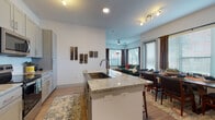This well-appointed single-floor residence offers a thoughtfully designed open-concept layout that seamlessly connects the kitchen, dining, and living areas. The kitchen is a standout feature, equipped with stainless steel appliances, a subway tile backsplash, granite countertops, and grey shaker-style cabinetry. A central island with a built-in sink provides additional prep space and doubles as a casual breakfast bar with seating. Light wood-style flooring flows throughout the main living areas, enhancing the bright, airy atmosphere created by multiple large windows dressed with blinds and floor-length drapes. The dining area accommodates a full-sized table and benefits from generous natural light. The living room features corner windows that invite the outdoors in, lending the space a relaxed, welcoming ambiance. Both bedrooms are comfortably sized, each with distinct character — one features a striking botanical wall mural, while the other includes a window with outdoor views. The two bathrooms are finished with matching granite vanities and grey cabinetry; one includes a tiled soaking tub and shower combo, while the other offers a glass-enclosed walk-in shower. The overall design balances modern finishes with warm, eclectic styling.