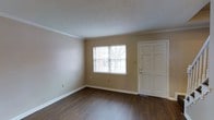 This two-floor residential space offers a clean and functional layout with classic design details that create a welcoming atmosphere. The entry area opens into a bright living space featuring rich, dark hardwood-style flooring that contrasts tastefully with the warm neutral beige walls. Crown molding runs along the ceiling line, adding a polished architectural touch to the room. A large window fitted with horizontal blinds allows natural light to filter in, enhancing the open and airy feel of the space. The textured ceiling adds subtle visual interest while maintaining a traditional aesthetic. White trim work and wide baseboards frame the room cleanly, complementing the six-panel white entry door. The overall palette of warm neutrals and dark flooring gives the main level a grounded, inviting character. Spanning two floors, the layout offers separation between living and private areas, providing both functionality and comfort. The design throughout emphasizes clean lines, traditional detailing, and a neutral color scheme that offers a versatile backdrop for a variety of interior styles.