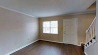 This two-floor residential space offers a clean and functional layout with classic design details that create a welcoming atmosphere. The entry area opens into a bright living space featuring rich, dark hardwood-style flooring that contrasts tastefully with the warm neutral beige walls. Crown molding runs along the ceiling line, adding a polished architectural touch to the room. A large window fitted with horizontal blinds allows natural light to filter in, enhancing the open and airy feel of the space. The textured ceiling adds subtle visual interest while maintaining a traditional aesthetic. White trim work and wide baseboards frame the room cleanly, complementing the six-panel white entry door. The overall palette of warm neutrals and dark flooring gives the main level a grounded, inviting character. Spanning two floors, the layout offers separation between living and private areas, providing both functionality and comfort. The design throughout emphasizes clean lines, traditional detailing, and a neutral color scheme that offers a versatile backdrop for a variety of interior styles.