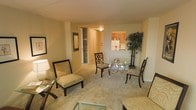 This single-floor residence presents an open-concept layout that seamlessly connects the living, dining, and kitchen areas. The living space is finished with plush wall-to-wall carpeting in a neutral tone, contributing to a warm and inviting ambiance throughout. Walls are painted in soft beige, enhancing the sense of light and continuity across the space.

The dining area features a round glass-top table accompanied by dark wood lattice-back chairs, offering an elegant yet understated focal point. A tall decorative bamboo plant adds a natural, organic element to the corner of the room. The living area is furnished with patterned armchairs and complementary accent tables, creating a comfortable and cohesive seating arrangement.

A hallway visible from the main living space leads to additional rooms and includes a built-in storage area with shelving. The kitchen, partially visible through an open passthrough, features warm wood cabinetry, light countertops, and standard appliances, providing a functional and well-organized cooking space. Overall, the residence offers a practical and comfortably appointed interior with a consistent warm, neutral aesthetic throughout.