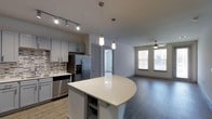 This well-appointed 2-bedroom, 2-bathroom single-floor residence offers a thoughtfully designed open-concept layout that seamlessly connects the kitchen, dining, and living areas. The modern kitchen is a standout feature, showcasing gray shaker-style cabinetry, a mosaic tile backsplash, and a full suite of stainless steel appliances including a French door refrigerator with ice maker and a dishwasher. A curved kitchen island with a quartz countertop provides additional prep space and casual seating, while pendant lighting above adds a stylish touch. Track lighting illuminates the kitchen workspace effectively. The open living area is finished with rich hardwood-style flooring and features a ceiling fan, creating a comfortable and functional space. A large window floods the living area with natural light and offers views to the outside. The neutral color palette throughout — soft grays and warm whites — lends a clean, contemporary feel. Interior doors feature classic panel detailing, adding architectural character. The overall ambiance is modern and inviting, balancing practical design with refined finishes that make this a comfortable and attractive living space.