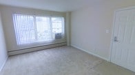 This single-floor space offers a clean, straightforward layout designed for comfortable, everyday living. The main room is a standout feature, characterized by large floor-to-ceiling windows dressed with vertical blinds that allow an abundance of natural light to fill the interior, creating a bright and airy atmosphere. The view through the windows reveals surrounding greenery, adding a pleasant outdoor connection to the space. Underfoot, plush wall-to-wall carpeting in a neutral beige tone provides warmth and comfort throughout the room. A baseboard heating unit runs along the base of the windows, complemented by a wall-mounted air conditioning unit, ensuring year-round climate control. The walls are finished in a soft, neutral tone that enhances the sense of openness and provides a versatile backdrop. White baseboards frame the room cleanly, contributing to the space's tidy, well-maintained appearance. Overall, the space presents a simple yet functional environment, well-lit and thoughtfully appointed with practical amenities that make it a comfortable and adaptable living area.