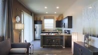 This single-story space showcases an open-concept layout that seamlessly connects the kitchen and living area, creating a cohesive and functional environment. The kitchen is a standout feature, designed with dark espresso cabinetry that extends to the upper walls, providing ample storage throughout. Granite countertops in warm, neutral tones complement the cabinetry and add a touch of elegance to the space.

A central kitchen island with matching granite surfaces doubles as a breakfast bar, complete with two industrial-style adjustable barstools. Stainless steel appliances, including a French door refrigerator with a water dispenser and a built-in microwave, enhance the modern aesthetic. Recessed lighting in the ceiling ensures the kitchen is well-lit, while a window above the sink allows natural light to filter in.

The wood-look flooring runs throughout, providing continuity between the kitchen and the adjoining living area. Large vertical blinds along the living area wall suggest floor-to-ceiling windows or sliding doors that bring in additional natural light. Decorative accents, including woven baskets and warm-toned pottery displayed atop the cabinetry, add character and warmth to the overall ambiance.