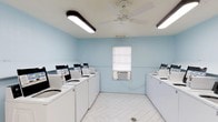 This shared laundry facility is designed with functionality and comfort in mind. The room features light blue walls that create a calm, clean atmosphere, complemented by white herringbone-patterned tile flooring that adds a subtle decorative touch while remaining practical and easy to maintain.

Multiple top-load washing machines are arranged along two walls, each equipped with digital control panels, providing ample capacity for simultaneous use. Overhead, two elongated fluorescent light fixtures ensure the space is brightly and evenly illuminated, while a white ceiling fan with an integrated light promotes air circulation throughout the room.

A wall-mounted air conditioning unit positioned beneath a frosted window ensures comfortable temperatures year-round, regardless of heat generated by the machines in use. The window also allows a degree of natural light into the space. The overall layout is open and spacious, allowing users to move freely between machines without congestion. The combination of practical amenities, efficient lighting, climate control, and a clean aesthetic makes this a well-appointed and user-friendly laundry facility.
