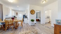 This single-floor home offers a well-organized layout designed for comfortable, everyday living. Light wood-look flooring runs consistently throughout the space, providing a cohesive and clean aesthetic that ties each area together seamlessly.

The galley-style kitchen is efficiently designed, featuring white cabinetry with dark countertops, stainless steel appliances including a refrigerator, range, and dishwasher, and a exterior-facing door that allows natural light to brighten the workspace. The kitchen flows directly into a dedicated dining area, which accommodates a round wooden table and chairs, creating a practical and inviting spot for meals.

A central hallway connects the main living areas, accented by a large decorative wall clock and a potted plant that add warmth to the transition space. The living room offers a comfortable arrangement with a sofa, armchair, and a leather recliner positioned around a wooden storage trunk used as a coffee table. A window fitted with curtains and blinds provides natural light to the room.

The bedroom is a calm, well-lit retreat featuring a window with grey curtains, a slatted wood-frame bed, a storage chest, and a wide dresser. The bathroom combines functional amenities with a vanity, toilet, walk-in shower, and in-unit washer and dryer, maximizing utility within a compact footprint.