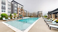 Located at 2551 Frontier Dr in Pittsburgh, PA, this outdoor amenity complex is designed to offer a comprehensive, resort-style living experience. At its centerpiece sits a generously sized swimming pool with crystal-clear blue water, surrounded by a broad pool deck lined with white lounge chairs and teal-accented umbrellas, providing ample space for relaxation and recreation. Adjacent to the pool is a covered pavilion featuring brick columns, ceiling fans, and large windows that overlook the pool area, creating a shaded retreat ideal for year-round use. The landscaped courtyards throughout the complex are thoughtfully designed with a mix of ornamental trees, flowering shrubs, and mulched garden beds, offering vibrant greenery and seasonal color. Wooden pergolas with climbing vines provide shaded gathering areas furnished with dining tables and chairs, perfect for outdoor entertaining. A dedicated sports court accommodates pickleball or tennis, surrounded by manicured plantings that add a polished, finished look. Concrete pathways connect each zone seamlessly, while Adirondack-style wooden chairs and benches are placed throughout for casual seating. The multi-story residential buildings framing the complex add architectural character with contrasting dark gray, brick, and light panel facades, creating a cohesive and modern community aesthetic.