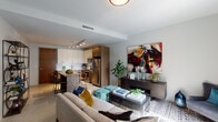 This 1-bedroom, 1-bathroom residence features a thoughtfully designed open-concept floor plan that seamlessly connects the kitchen, dining, and living areas. The contemporary kitchen is outfitted with stainless steel appliances — including a refrigerator, electric range, and over-the-range microwave — complemented by two-tone cabinetry featuring white upper cabinets and warm wood-toned lower cabinets, white quartz countertops, and a classic subway tile backsplash. A built-in kitchen island with bar seating provides additional prep space and a casual dining option. The open living area offers generous room for lounging and entertaining, filled with natural light streaming through floor-to-ceiling windows dressed with teal drapery. The bedroom is a serene retreat with large windows framing outdoor views, a ceiling fan, and ample space for furnishings. The full bathroom is finished with large-format marble-look tile surrounding the tub and shower, a wood-toned vanity with quartz countertop, and polished concrete-style flooring. Track lighting throughout the main living areas adds a modern touch, while the cohesive neutral palette creates a clean, inviting atmosphere throughout.