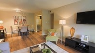This single-story space showcases an open-concept floor plan that seamlessly connects the living, dining, and kitchen areas, creating a cohesive and functional layout. Warm hardwood flooring extends throughout the main living areas, adding a sense of continuity and warmth to the interior.

The living area is defined by neutral, beige-toned walls that provide a versatile backdrop for furnishings and décor. A tufted accent chair in sage green adds a pop of color, complemented by coordinating orange and patterned throw pillows. A glass-top coffee table anchors the seating arrangement, while a sleek media console and floor lamp contribute to the room's layered lighting design.

Adjacent to the living space, the dining area accommodates a compact table and chairs, illuminated by a track lighting fixture overhead. Framed artwork along the wall adds visual interest to this corner of the space.

The kitchen is partially visible through an open passageway, where stainless steel appliances, including a full-size refrigerator, can be seen. The overall ambiance is warm and inviting, characterized by clean lines, a neutral color palette, and a practical, well-connected layout suited for everyday living.