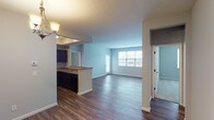 This single-floor residence offers a thoughtfully designed open-concept layout that seamlessly connects the living, dining, and kitchen areas. Rich, dark hardwood-style flooring flows throughout the main living spaces, complemented by neutral light-gray walls that create a clean, modern aesthetic. The dining area features an elegant chandelier, while large windows at the front of the living area allow natural light to fill the space.

The kitchen is equipped with dark espresso cabinetry, granite-style countertops, black appliances including a range and dishwasher, and a functional peninsula with a sink that opens to the dining area — ideal for both cooking and entertaining.

The bedrooms are carpeted for comfort, each featuring neutral gray walls, natural window light, and panel-style doors. A spacious walk-in closet with wire shelving and hanging rods provides generous storage. Two full bathrooms mirror each other in design, each featuring tile-surround soaking tubs, checkered tile flooring, dark vanity cabinetry, and white countertops. The layout, as seen in the dollhouse view, offers a well-organized flow between the public and private areas of the home, making efficient use of the available space throughout.
