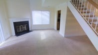 This two-story home features a spacious main living area finished with plush, light-toned carpeting that extends throughout the floor. The living room serves as the central gathering space, highlighted by a classic fireplace with a dark surround and hearth, adding both a functional and aesthetic focal point to the room. A large window fitted with horizontal blinds allows natural light to fill the space, brightening the neutral, cream-toned walls.

A staircase situated in the corner of the living area provides access to the upper floor, its white balusters and railing complementing the clean, understated interior palette. Adjacent to the living room, an open passageway leads into the kitchen, where warm wood-toned cabinetry is visible, offering a glimpse of the cooking and preparation space beyond.

The overall design is straightforward and practical, with high walls that contribute to an open, airy feel. The neutral color scheme throughout creates a versatile backdrop suitable for a variety of interior styles. The layout efficiently connects the main living space to both the kitchen and the upper level.