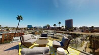 This rooftop terrace is defined by its sweeping, unobstructed views of a Southern California cityscape, with palm trees and mid-rise buildings stretching across the horizon beneath consistently clear blue skies. The outdoor deck is finished with warm wood-look porcelain tiles laid in a herringbone pattern, lending a polished yet inviting aesthetic throughout the space.

The terrace is organized into several distinct seating zones. One area features woven bistro-style chairs and round mosaic tables, ideal for casual dining or social gatherings. Adjacent to this, a lounge area is furnished with cushioned sofas and armchairs arranged on a bold yellow geometric area rug, creating a relaxed, residential feel. A third zone near the building's exterior wall offers a sectional sofa set on a patterned rug, providing a more sheltered retreat.

A built-in outdoor kitchen with a gas grill and planter boxes filled with succulents lines one wall, adding functionality and a touch of greenery. Glass panel railings line the perimeter, preserving sightlines while ensuring safety. Access to the rooftop is provided via an elevator lobby finished with patterned carpet tiles and recessed lighting, offering a clean, modern transition from the building's interior to the expansive outdoor deck above.