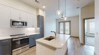 Located at 4501 E Warren Ave in Denver, Colorado, this single-floor residence offers a thoughtfully designed layout with modern finishes throughout. The open-concept kitchen serves as the centerpiece of the living area, anchored by a large island with a white quartz countertop and an undermount sink. Two-tone cabinetry — pairing white upper cabinets with slate-gray lower cabinets — lines the walls alongside a classic white subway tile backsplash. Stainless steel appliances, including a refrigerator, range, microwave, and dishwasher, complete the well-appointed kitchen. Globe pendant lights hang above the island, adding a contemporary touch, while light wood-look flooring runs throughout the main living area. A bold teal entry door makes a striking first impression. The bedroom is carpeted and bathed in natural light from a large window with exterior views. The bathroom features wood-look tile flooring, a dual-sink vanity with white countertops and gray cabinetry, a framed mirror, and a tiled walk-in shower. Panel-style gray interior doors carry a cohesive design language across the unit. The living area opens to a windowed door, allowing additional natural light to filter in and connecting the interior to the outdoors.