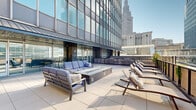 Located at 55 Public Square in the heart of downtown Cleveland, this generously sized amenity floor offers a sophisticated blend of interior comfort and outdoor urban living. The open-concept interior lounge is designed for both relaxation and social gatherings, featuring tray ceilings with elegant gold chandelier fixtures, recessed lighting, and a sleek linear fireplace set against a richly textured dark tile surround. Seating areas are thoughtfully arranged throughout, with plush velvet chairs, leather sofas, café-style tables, and a full shuffleboard table for recreation. A wall-mounted flat-screen TV anchors one end of the lounge.

The fully equipped catering kitchen is a standout feature, outfitted with white shaker-style cabinetry, a bold blue subway tile backsplash, a large center island with a white quartz countertop, stainless steel appliances including double wall ovens and a full-size refrigerator, and globe pendant lighting overhead.

Step outside to the expansive rooftop terrace, where large-format tile pavers create a clean, modern surface. A central fire pit table is surrounded by cushioned lounge seating and chaise lounges, all set against sweeping views of the Cleveland skyline, including the iconic Terminal Tower. Bar-height seating and greenery complete this impressive outdoor retreat.
