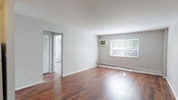 This two-bedroom, single-floor unit offers a clean and functional layout designed for comfortable everyday living. The open living area is finished with rich, dark hardwood-style flooring that adds warmth to the neutral gray walls, creating a bright and inviting atmosphere. A large window allows generous natural light to fill the space, with views of greenery outside. The unit is equipped with baseboard heating and a wall-mounted air conditioning unit for year-round comfort.

The kitchen features dark cabinetry contrasted against white appliances, including an electric range and refrigerator, along with a sink and ample cabinet storage. Light wood-style flooring ties the kitchen into the rest of the unit seamlessly.

Both bedrooms are well-proportioned, each with hardwood-style flooring, baseboard heaters, and windows that bring in natural light and outdoor views. One bedroom includes a wall-mounted air conditioning unit for added convenience. Closet storage is also present, as seen from the dollhouse view.

The bathroom is neatly appointed with a tub and shower combination featuring a grab bar, a vanity with a light countertop, and tile flooring. The overall design of this unit is straightforward and practical, offering a comfortable and move-in ready living space.