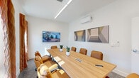 Located at 14375 S Maplelane Rd in Oregon City, Oregon, this single-floor conference room offers a polished and professional meeting environment. The space is anchored by a substantial light oak boardroom table surrounded by tan leather executive chairs, comfortably accommodating groups for collaborative sessions or formal meetings. Crisp white walls and a neutral carpet create a clean, contemporary backdrop, while a pair of geometric abstract wall art panels adds a refined decorative touch. A wall-mounted flat-screen display provides integrated presentation capabilities, and a mini-split climate control unit ensures year-round comfort. Recessed linear lighting overhead delivers consistent, bright illumination throughout. Large exterior windows dressed with warm-toned curtains invite generous natural light and offer views of the surrounding outdoor environment. Frosted glass interior doors and a glass partition wall connect the room to the adjacent areas while preserving privacy, contributing to a bright and open feel. Potted snake plants placed along the table introduce a subtle organic element to the otherwise modern aesthetic. The overall ambiance is clean, functional, and thoughtfully appointed, making it well-suited for professional gatherings, presentations, or business meetings.
