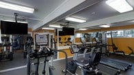 This fitness center is designed to provide a functional and motivating workout environment. The space is outfitted with a comprehensive selection of TRUE-brand cardio equipment, including elliptical trainers, upright stationary bikes, and treadmills, all arranged to maximize accessibility and floor space. Overhead fluorescent lighting ensures the room is well-lit throughout, contributing to a bright and energetic atmosphere. Bold orange accent walls add a dynamic visual element, complementing the neutral gray flooring and white structural beams that define the ceiling. A wall-mounted flat-screen television is centrally positioned, offering entertainment during workouts. Full-length mirrors line one wall, visually expanding the space while allowing users to monitor their form. A windowed wall or door provides a view to the exterior, introducing a degree of natural light. The overall layout is practical and organized, with clear pathways between equipment. The space reflects a straightforward, purpose-driven design focused on delivering essential fitness amenities in a clean, accessible setting.