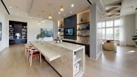 This elegantly designed single-floor space makes a strong first impression with its open, airy layout and refined architectural details. Light-toned hardwood flooring flows seamlessly throughout, lending warmth and continuity to the entire space. Striking black-framed glass French doors serve as a focal point near the entry, offering a glimpse into adjoining areas while allowing natural light to pour in from large windows that frame outdoor views.

The central gathering area is anchored by a substantial marble-topped island with integrated shelving below, doubling as both a functional surface and a design statement. A long communal dining table, accented by warm amber glass pendant lights, sits adjacent to a dramatic built-in media wall featuring backlit open shelving, dark cabinetry, and decorative tilework that adds texture and visual depth.

Curated contemporary artwork is thoughtfully displayed across the crisp white walls, including a bold abstract canvas in deep blues and golds that draws the eye and elevates the cultural atmosphere of the space. A compact accent console in red with gold legs adds a playful pop of color near the entryway, complemented by a gold-framed mirror above.

The overall ambiance is modern yet inviting, blending clean architectural lines with eclectic decorative touches to create a space that feels both polished and welcoming.