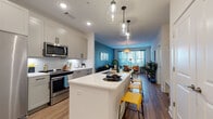 Located at 2200 Silver Crescent Dr in Charlotte, NC, this single-floor residence offers a thoughtfully designed open-concept layout that seamlessly connects the kitchen, dining, and living areas. The kitchen is a standout feature, showcasing gray shaker-style cabinetry with matte black hardware, a herringbone tile backsplash, white quartz countertops, and a full suite of stainless steel appliances including a refrigerator, range, microwave, and dishwasher. A central island with an undermount sink and pendant lighting above serves as both a functional workspace and casual dining spot. Rich wood-look flooring flows throughout the main living areas, anchoring the open and airy atmosphere. The living room is characterized by a bold teal accent wall, large windows that invite abundant natural light, and direct access to an outdoor view. The bedroom continues the vibrant aesthetic with a warm yellow accent wall, plush carpeting, and generous windows that brighten the space. The bathroom is clean and functional, offering built-in open shelving and direct access to a spacious walk-in closet with wire shelving. Throughout the residence, the design balances modern finishes with bold color choices, creating a lively yet cohesive living environment.