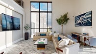 This contemporary amenity space is defined by its dramatic double-height ceilings and expansive floor-to-ceiling windows framed in sleek black steel, flooding the interiors with natural light and offering open views of the surrounding streetscape and skyline. The entry welcomes visitors through a bright, glass-enclosed vestibule finished with large-format marble-look tile flooring that continues throughout the main areas.

The open-plan layout connects several distinct zones seamlessly. A sophisticated lounge area features neutral-toned sectional sofas, patterned area rugs, and tasteful greenery, anchored by a wall-mounted display screen. An upper-level mezzanine with ornamental iron railings overlooks the main floor, adding architectural depth.

The kitchen and dining area is a standout, showcasing bold navy-blue cabinetry, open wood-accented shelving, a dark stone island with bar seating, stainless steel appliances, and a decorative linear pendant light. A marble-top round dining table with powder-blue upholstered chairs adds an elegant touch.

A vibrant game room features a teal accent ceiling, a full-size pool table, lounge seating, and a large mounted television. Large windows in this area provide additional street-level views, enhancing the open, airy atmosphere throughout the space.
