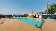 This outdoor pool area offers a clean, functional, and inviting retreat open to the sky. The centerpiece is a generously sized rectangular swimming pool with clearly marked depth indicators ranging from a shallow entry point to a deeper end, making it accessible for swimmers of all skill levels. The pool's calm, turquoise water is complemented by a broad concrete deck that wraps around all sides, providing ample space for lounging and movement. Scattered across the deck are lightweight chairs and vibrant teal Adirondack-style seating, offering spots to relax in the open air. The pool is accessed via stainless steel handrails and entry steps, ensuring safe and convenient entry and exit. The entire area is enclosed by a combination of wooden privacy fencing and a metal safety fence, creating a secure and contained environment. Posted pool rules signage is prominently displayed near the entry, reinforcing safe usage guidelines. The surrounding landscape includes glimpses of palm trees and open sky, lending a warm, sun-drenched atmosphere characteristic of a southern California setting. The adjacent building structure is visible from within the pool area, grounding this amenity space within a larger property complex.