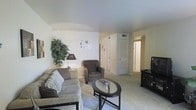 This single-floor residential space offers a warm and functional layout designed for everyday comfort. The living room serves as the heart of the home, featuring plush seating including a cushioned sofa adorned with accent pillows and a matching upholstered armchair, arranged around a sleek glass-top coffee table. Soft, neutral-toned carpeting runs throughout, contributing to the room's cozy and inviting ambiance. Decorative wall accents and a lush indoor plant add a touch of personality and life to the space. A floor lamp provides warm accent lighting, complementing the room's understated aesthetic. The living area flows naturally toward a hallway that connects to additional rooms, with doors visible along the corridor suggesting well-defined private spaces beyond. An entertainment setup is positioned along one wall, offering a dedicated area for relaxation. The ceilings are smooth and finished in white, enhancing the sense of openness. Ventilation is provided via ceiling-mounted HVAC vents, ensuring year-round comfort. Overall, the space presents a clean, classic interior with a practical and livable design that balances simplicity with functionality.