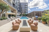 Located at 3775 Sixth Avenue in San Diego's vibrant Hillcrest neighborhood, this generously scaled amenity level delivers a resort-caliber outdoor and indoor experience across nearly 5,000 square feet. At its centerpiece is a long, sparkling lap pool flanked by rows of sun loungers shaded by large umbrellas, set against a backdrop of a striking multi-story residential tower. Along one side, a covered colonnade lined with lush potted palms and tropical plantings provides a shaded walkway connecting the various zones of the deck. Adjacent to the pool, a slatted wooden pergola shelters a collection of plush cabanas and lounge furniture, creating intimate seating areas ideal for relaxed outdoor gatherings. A semi-covered outdoor kitchen and entertaining terrace features warm stucco walls, wood cabinetry, a prep counter, and open-air dining nooks surrounded by mature trees and curated landscaping. The interior clubroom commands attention with soaring double-height ceilings, floor-to-ceiling black-framed glass walls that flood the space with natural light, and a dramatic central fireplace. Woven pendant lighting, built-in banquette seating, and layered textiles in earthy tones lend a sophisticated, spa-like ambiance throughout. A terrace lounge with fire pit seating and decorative screen panels seamlessly bridges the indoor and outdoor areas, completing this thoughtfully designed communal retreat.