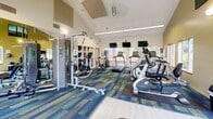Located at 11540 Apache Dr in Parma Heights, Ohio, this dedicated fitness center offers a well-equipped exercise space designed to support a comprehensive workout routine. The room features striking vaulted ceilings that lend an open, airy feel, complemented by warm yellow-green accent walls and a vibrant multi-toned blue and green patterned carpet that adds energy to the environment. Large windows along multiple walls flood the space with natural light and provide pleasant views of the surrounding greenery, creating an inviting atmosphere for exercise. The cardio section is equipped with multiple Matrix treadmills and elliptical trainers, each featuring individual digital displays, as well as a recumbent stationary bike. Wall-mounted flat-screen televisions are positioned for easy viewing from the cardio equipment. The strength training area includes a multi-station cable machine, a weight bench, stability balls, and a full rack of dumbbells, catering to a wide range of fitness needs. A large wall mirror enhances the sense of space and allows for proper form checks during workouts. Built-in display shelving adds a decorative architectural touch to one wall. The flooring transitions between cushioned carpet and hard surface areas, providing appropriate surfaces for both equipment use and functional training. The overall ambiance is bright, functional, and well-maintained.