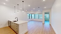 This single-floor residence offers a clean, contemporary design defined by an open-concept layout and an abundance of natural light. Light wood flooring runs continuously throughout the living areas, complementing the crisp white walls and recessed lighting overhead. Large floor-to-ceiling windows frame sweeping panoramic views of a high-rise urban skyline, bringing the city's energy directly into the living and bedroom spaces.

The open kitchen is thoughtfully appointed with a central island featuring a white quartz countertop, an undermount sink with a matte black faucet, and pendant lighting above. Sage-toned cabinetry with black hardware houses ample storage, while stainless steel appliances — including a built-in microwave and wine refrigerator — add a polished, functional touch.

The primary bedroom enjoys its own dramatic city views through expansive windows, creating a bright and airy retreat. A spacious walk-in closet features built-in shelving and double hanging rods, offering generous organization capacity. An additional closet provides further storage.

The bathroom is finished with warm wood cabinetry, a white quartz vanity, a backlit mirror, and a glass-enclosed walk-in shower with tile surround and matte black fixtures — delivering a spa-like aesthetic. The overall ambiance of the space is light, airy, and refined, ideal for modern urban living.