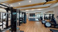Located at 3529 Adirondack Way in Wilmington, NC, this single-story fitness facility offers a comprehensive workout environment designed with both function and aesthetics in mind. The space features a distinctive modern farmhouse interior, highlighted by white shiplap walls, warm wood beam accents across the ceilings, and durable light-toned hardwood flooring throughout.

The main workout area is generously proportioned, housing a full complement of professional-grade equipment including a Precor cable machine system, a heavy punching bag, a multi-functional power rack with integrated storage for dumbbells and accessories, and TRX suspension trainers. A dedicated cardio zone features multiple treadmills, elliptical trainers, and a rowing machine, all positioned along large windows that invite abundant natural light and outdoor views.

Additional training areas include a strength-focused nook with an adjustable bench and specialized equipment, offering a more intimate setting for focused workouts. Floor-to-ceiling mirrors are strategically placed throughout to enhance the sense of space and support proper form. A built-in wet bar area with slate-blue cabinetry provides convenient access to hydration. The entryway features stylish slate-blue double doors, recessed lighting, and built-in wooden shelving, creating a polished and welcoming transition into the facility. Ceiling fans maintain comfortable airflow throughout the space.