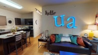 This single-floor residential unit offers a thoughtfully designed open-concept layout that seamlessly connects the living, dining, and kitchen areas. The living room is a welcoming space anchored by warm hardwood-style flooring, a comfortable sofa, and a dining area with bar-height seating — ideal for both relaxing and entertaining.

The kitchen is a standout feature, outfitted with rich dark espresso cabinetry, granite countertops, a full suite of black appliances including a refrigerator, electric range, microwave, and dishwasher, plus a convenient kitchen island for additional workspace and storage. Track lighting overhead keeps the space bright and functional.

The unit includes multiple private bedrooms, each carpeted for comfort and featuring neutral tones, ample wall space, and natural light from windows with blinds. The bedrooms offer a calm, restful atmosphere suited for study or sleep.

Each full bathroom is finished with dark wood cabinetry, granite-style countertops, hardwood-style flooring, and a tub-shower combination — providing a clean and cohesive aesthetic throughout. The layout of the unit, as revealed in the dollhouse view, accommodates multiple residents comfortably, with clear separation between shared and private spaces.