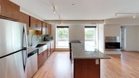 This well-appointed single-floor unit offers a thoughtfully designed open-concept layout that seamlessly connects the kitchen and living areas. The kitchen is a standout feature, showcasing rich warm-toned wood cabinetry, granite countertop surfaces, and a large central island with an integrated cooktop — ideal for both meal preparation and casual dining. Stainless steel appliances, a mosaic tile backsplash, and track lighting overhead add a contemporary, polished feel. Multiple large windows flood the kitchen and living space with natural light and offer pleasant outdoor views of surrounding greenery. The unit includes two bathrooms: one featuring a walk-in glass-enclosed shower with chrome fixtures and a granite-topped vanity, and a second full bathroom with a soaking tub/shower combination and matching granite vanity. Both bathrooms are finished with neutral tile flooring and clean white walls, maintaining a bright and functional aesthetic. The bedroom features plush wall-to-wall carpeting and a sliding closet, providing a comfortable and quiet retreat. Light hardwood flooring runs throughout the main living and kitchen areas, tying the space together with warmth and continuity. Overall, the unit presents a clean, modern design with quality finishes throughout.