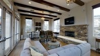 This single-story space showcases a thoughtfully designed interior that blends rustic and contemporary elements. The living area is immediately distinguished by its striking exposed dark wood ceiling beams, which add architectural character and visual depth. Recessed lighting and two elegant chandelier-style pendant fixtures illuminate the space, complementing the natural light that streams in through multiple windows framed with dark valances.

The stacked stone fireplace serves as a commanding focal point along one wall, paired with a wall-mounted flat-screen television above, combining classic and modern design sensibilities. The hearth extends with a clean white surround, providing a refined contrast to the textured stonework.

Warm hardwood floors run throughout, anchoring a comfortable seating arrangement that includes a large tufted sectional sofa, wingback accent chairs, and a rustic round coffee table. A cozy bistro-style dining nook with bar-height seating and decorative wall shelving occupies one corner, offering a casual space for smaller gatherings.

The neutral color palette of warm beiges and taupes, accented with soft blue decorative pillows and natural greenery, creates a welcoming and cohesive ambiance throughout the space.
