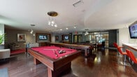 This single-floor game and recreation lounge is designed as a comprehensive social and entertainment amenity. The space is anchored by multiple full-sized billiard tables featuring vibrant red felt surfaces, complemented by a long shuffleboard table and a foosball table, providing a well-rounded selection of game options. Rich, dark hardwood flooring runs throughout the main area, lending warmth and a polished aesthetic to the room.

Overhead, distinctive pendant light fixtures with teardrop-shaped glass globes hang from the ceiling alongside adjustable track lighting, ensuring the gaming areas are well-illuminated. Multiple wall-mounted flat-screen televisions are positioned throughout the space, making it suitable for watching sports or entertainment alongside gameplay.

A lounging area with contemporary swivel chairs and red accent rugs provides comfortable seating for spectators or those taking a break from the games. A wet bar or refreshment counter with bar seating is visible toward the rear of the space, enhancing its function as a social gathering area.

Large glass doors and windows along one wall allow natural light to filter in and provide views of the outdoor surroundings. The overall design balances an energetic, activity-oriented atmosphere with modern, upscale finishes, making it a versatile and inviting communal space.