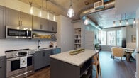 This single-floor unit showcases an urban industrial aesthetic defined by exposed concrete ceilings and visible ductwork throughout. The open-concept kitchen is a standout, featuring gray shaker cabinetry, stainless steel appliances, a white subway tile backsplash, a matte black faucet, and a generously sized island with a marble-veined quartz countertop and built-in shelving below. Warm Edison bulb pendant lighting adds an inviting glow above both the island and the adjoining dining area. Wood-tone flooring runs throughout, complementing the raw industrial elements overhead.

The living area features a painted brick accent wall, large windows that draw in natural light, and an open sightline to the kitchen. Two bedrooms offer a tranquil retreat, each with floor-to-ceiling windows, exposed concrete ceilings, and neutral tones. One bedroom features a bold navy accent wall, while the other is finished in soft gray. The bathroom is appointed with a double-sink vanity, an illuminated mirror, hardwood flooring, and direct access to a well-organized walk-in closet. The overall ambiance balances industrial character with contemporary comfort.
