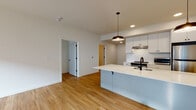 Located at 7900 SE Luther Rd in Portland, Oregon, this single-floor residence offers a thoughtfully designed open-concept layout with clean, modern finishes throughout. Warm hardwood-style flooring flows seamlessly from room to room, creating a cohesive and inviting atmosphere.

The kitchen serves as a central highlight, featuring a large peninsula with a white quartz countertop, undermount sink with a matte black faucet, and stylish pendant lighting overhead. White shaker-style cabinetry with black hardware pairs with a textured tile backsplash, while stainless steel appliances — including a range, refrigerator, and microwave — complete the functional and contemporary cooking space.

The open living area is bright and airy, benefiting from natural light admitted through large windows that offer views of the surrounding neighborhood. Recessed lighting and additional pendant fixtures enhance the well-lit interior.

The bathroom features a dark-cabinetry vanity topped with a white quartz surface, an undermount sink, and a large mirror with stylish overhead lighting. In-unit laundry is conveniently located nearby, adding practical day-to-day functionality.

The bedroom is generously proportioned, filled with natural light from oversized windows, and finished with the same consistent hardwood-style flooring. The overall design aesthetic is clean, modern, and move-in ready.