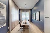 Located at 2926 Barker Cypress Rd in Houston, TX, this compact yet well-appointed business center offers a thoughtfully designed workspace within a single-floor layout. The room is defined by bold navy blue accent walls that contrast elegantly against the white ceiling and light-toned large-format tile flooring laid in a herringbone pattern, creating a polished and contemporary aesthetic.

At the heart of the space sits a long, light wood communal work table accommodating multiple workstations, each equipped with monitors and ergonomic rolling chairs. Natural light filters generously through floor-to-ceiling windows dressed in flowing sheer linen drapes, lending the room an airy and open atmosphere. Overhead, a striking angular pendant light fixture with linear LED elements serves as a focal point, complemented by recessed ceiling lighting throughout.

Framed artwork adorns the navy feature wall, adding a curated, professional touch. The space connects openly to an adjacent hallway, where a bank of parcel lockers and a glimpse of a communal kitchen or lounge area are visible, suggesting this business center is part of a larger amenity-rich facility. The overall ambiance is clean, modern, and functional — designed to support focus and productivity.