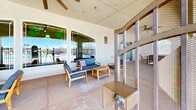 Located at 9800 Mesa Linda St in Hesperia, CA, this single-story community clubhouse offers a generously sized floor plan designed to support a variety of recreational and social activities. The expansive living room serves as the heart of the interior, featuring soaring ceilings, light wood-style flooring, and a striking floor-to-ceiling stone accent wall with a built-in fireplace and mounted television. Arched doorways connect the space to additional seating areas, while large windows flood the room with natural light and provide views of the surrounding outdoor amenities. A covered outdoor patio adjacent to the main lounge offers a comfortable seating area, perfect for relaxing while overlooking the sparkling freeform pool, which is surrounded by lounge chairs and patio seating. The dedicated fitness center is equipped with a multi-station weight machine, stationary bikes, a treadmill, and other cardio equipment, with windows providing natural light and views of the outdoor courts. Outside, a full-size basketball court enclosed by chain-link fencing offers scenic mountain views beyond its perimeter. A dedicated garden area features raised wooden planting beds set within a gravel landscape, complemented by a palm tree and open sky views. This well-rounded facility provides comprehensive amenities suited for an active residential community.