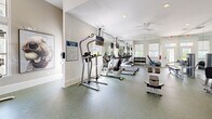 Located at 1575 Ridenour Parkway in Kennesaw, Georgia, this well-appointed fitness center offers a comprehensive exercise environment within a single, open floor plan. The space is thoughtfully designed with a clean, modern aesthetic, featuring neutral-toned walls, recessed lighting, and durable speckled rubber flooring that is both functional and visually appealing.

The exercise room is generously equipped to support a full range of workout routines. Cardio equipment includes multiple TRUE treadmills and elliptical machines arranged in a row, each outfitted with individual monitors for entertainment during workouts. Strength training is supported by a multi-station cable machine, a leg press, a weight bench, and a dumbbell rack, providing options for both resistance and free weight exercises.

Full-length wall mirrors run along one side of the room, amplifying the sense of space and allowing users to monitor their form. Ceiling fans provide comfortable air circulation, complementing the natural light that streams in through several windows and glass-paneled doors. Wall-mounted flat-screen televisions are positioned throughout the space for added convenience.

A posted fitness rules sign near the entrance helps maintain an orderly environment. The overall ambiance is bright, open, and purpose-built for an effective fitness experience.
