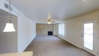 This single-floor residence offers a practical and comfortable layout with two bedrooms and two bathrooms. The living room serves as the heart of the home, highlighted by a classic full-height brick fireplace that adds warmth and character to the space. Built-in shelving flanks the fireplace, providing both display and storage options. A ceiling fan with integrated lighting sits centrally overhead, ensuring year-round comfort and even air circulation. The room is finished with neutral wall-to-wall carpeting that runs throughout, contributing to a cozy and unified aesthetic. A textured popcorn ceiling adds a traditional touch, while a large vertical-blind window allows natural light to filter into the space. The open, uncluttered floor plan offers ample room for a variety of furniture arrangements, making the living area both versatile and inviting. The overall ambiance of the home is warm and functional, combining classic design elements such as the exposed brick fireplace with practical features that suit everyday living. With two full bathrooms and two bedrooms complementing the spacious common areas, this residence provides a well-balanced and livable environment.
